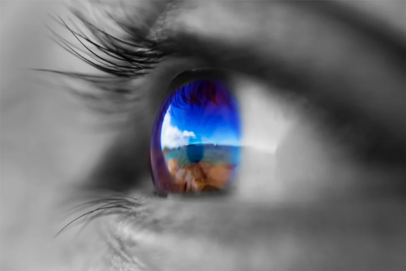 Kreatives Porträt/Makrofoto eines Auges, in dem eine Landschaft und der blaue Himmel reflektieren, vor einem unscharfen, schwarz-weißen Hintergrund.