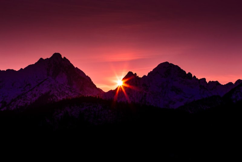 Foto eines dramatischen Sonnenaufgangs in den Alpen, bei dem die Sonne zwischen zwei schneebedeckten Berggipfeln aufgeht.