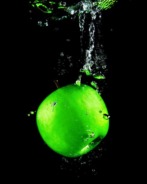 Ein grüner Apfel fällt ins Wasser, umgeben von präzise eingefangenen Wasserspritzern vor einem schwarzen Hintergrund.