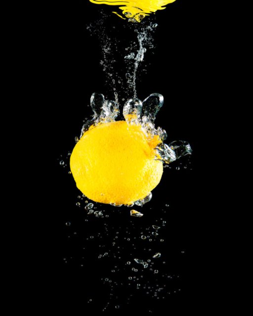 Eine gelbe Zitrone fällt ins Wasser, umgeben von präzise eingefangenen Wasserspritzern vor einem schwarzen Hintergrund.