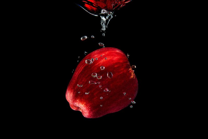Ein roter Apfel fällt ins Wasser, umgeben von präzise eingefangenen Wasserspritzern vor einem schwarzen Hintergrund.