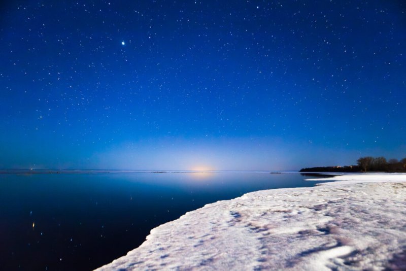 Foto eines Nachthimmels mit vielen Sternen über einem zugefrorenen See, mit einem schneebedeckten Ufer im Vordergrund.