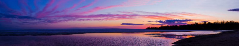 Panoramabild eines Strandes mit einem Sonnenuntergang, bei dem der Himmel in violetten, rosa und orangefarbenen Tönen leuchtet.