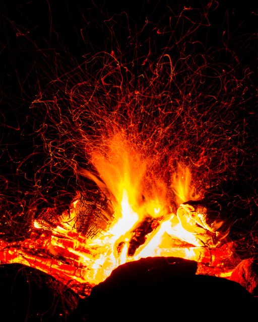Langzeitbelichtung eines Lagerfeuers mit hell leuchtenden Flammen und Funken vor einem dunklen Hintergrund.