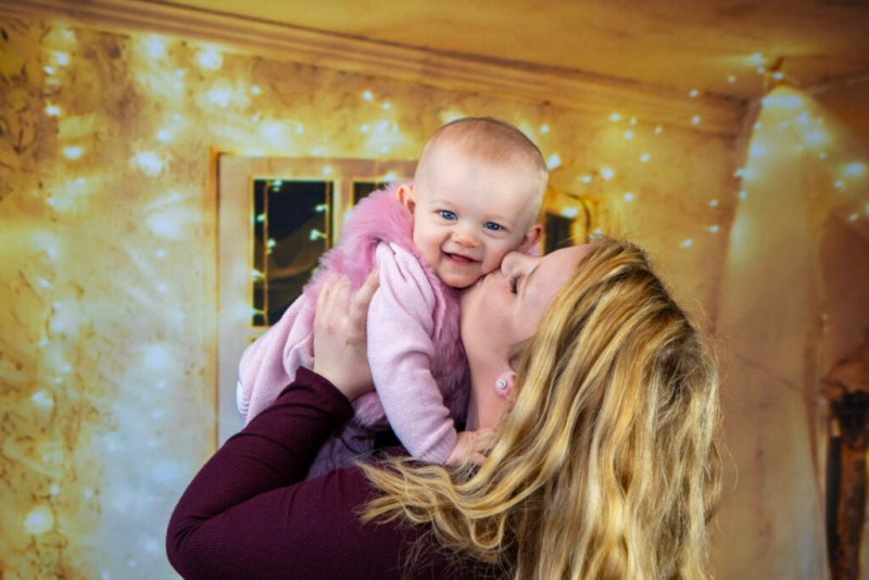 Eine junge Mutter küsst liebevoll ihr Baby, das in einem rosa Kleidchen gekleidet ist, vor einem hintergrundbeleuchteten goldenen Wanddekor.