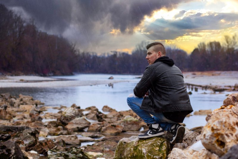 Ein junger Mann sitzt auf einem Felsen und blickt nachdenklich auf einen Fluss unter einem dramatischen Himmel.