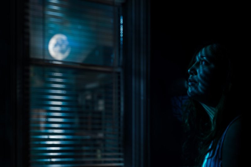 Eine Frau betrachtet nachdenklich durch ein Jalousiefenster, beleuchtet von blauem Neonlicht, das eine geheimnisvolle Stimmung schafft.