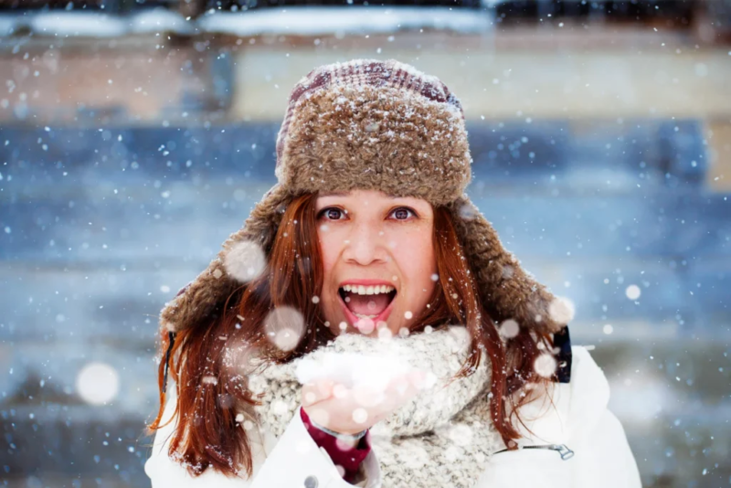 Branding-Porträt einer jungen Frau im verschneiten Winter, die in die Kamera lächelt. Sie trägt eine Wintermütze, Schal und Mantel.