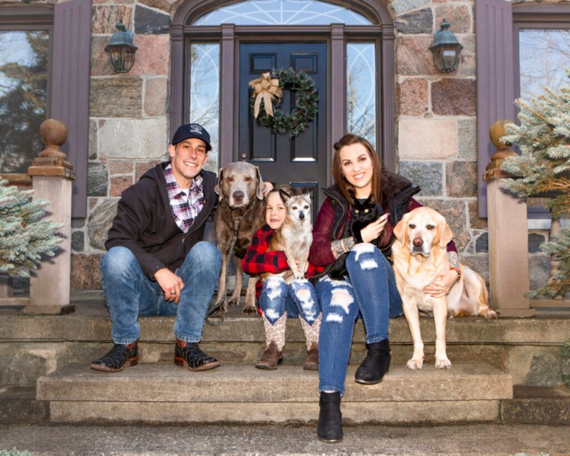 Eine glückliche Familie mit zwei Erwachsenen, einem Kind und vier Hunden sitzt auf der Haustreppe ihres Hauses, dekoriert mit Weihnachtsschmuck.
