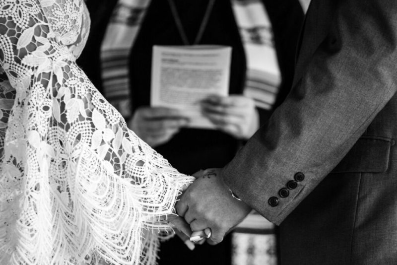 Schwarz-Weiß-Aufnahme der Hände eines Brautpaares, die sich während der Trauung halten, vor dem Hintergrund eines Pfarrers mit Traubuch.