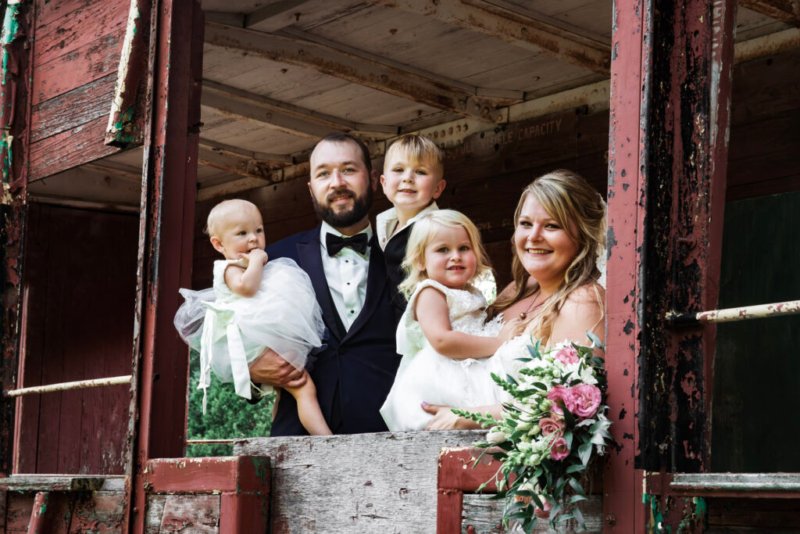 Junge Familie posiert fröhlich auf einem alten, rostigen Eisenbahnwaggon bei ihrer Hochzeitsfeier.