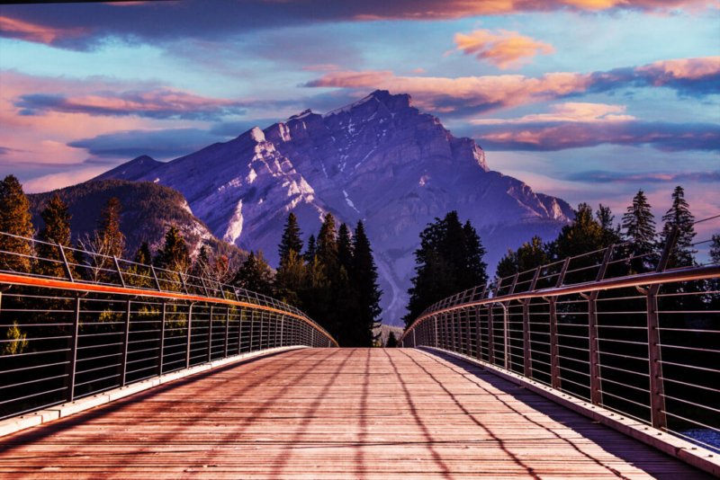 Foto der Brücke, die zum Cascade Mountain in Banff, Kanada, führt, umgeben von Bäumen und vor einem Himmel in warmen Farbtönen.