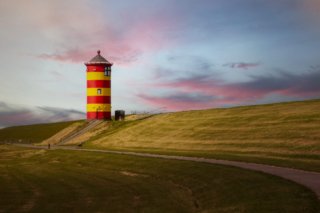 Foto des Pilsumer Leuchtturms mit roten und gelben Streifen auf einem grasbewachsenen Deich vor einem Himmel in rosa und blauen Farbtönen.