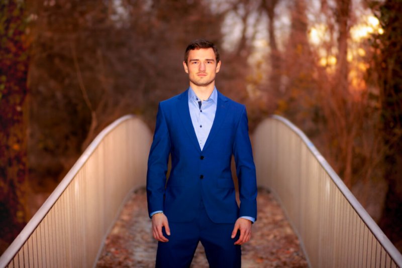 Junger Mann in elegantem blauen Anzug steht auf einer Brücke vor herbstlichem Hintergrund.