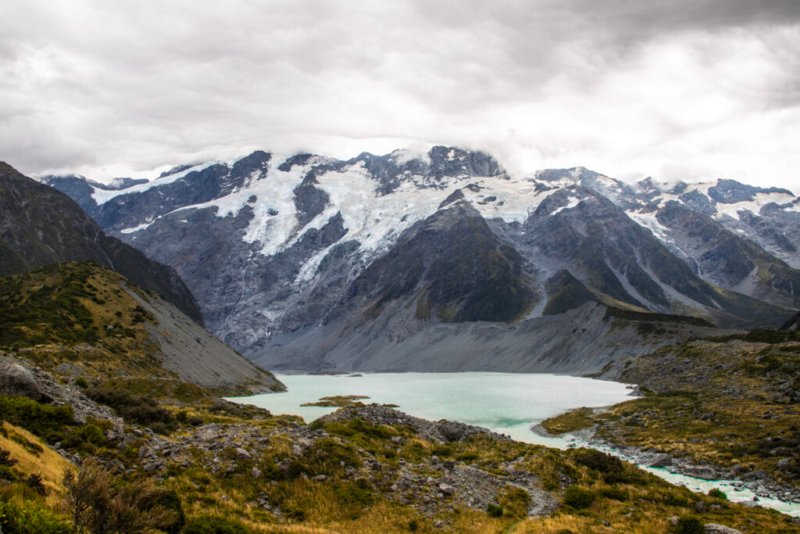 Foto des Franz-Josef-Gletschers in Neuseeland mit einem See und einer zerklüfteten Berglandschaft im Vordergrund.