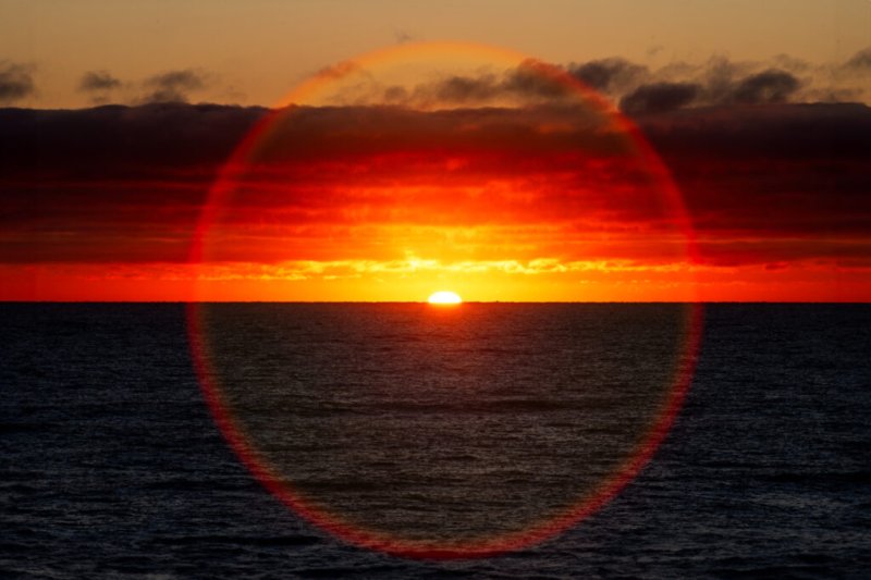 Foto eines leuchtenden Sonnenaufgangs über dem Meer mit rötlich-orangen Wolken und einem Lichtkreis im Vordergrund.