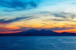 Foto des Lake Taupo in Neuseeland bei Sonnenuntergang, mit farbenprächtigen Wolken am Himmel und einer Bergkette im Hintergrund.