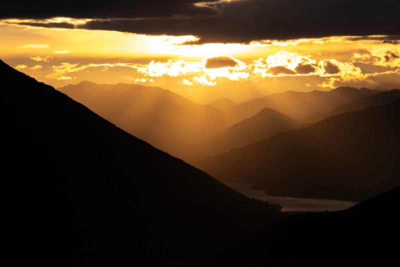 Landschaftsfoto eines Sonnenuntergangs hinter Bergketten, die im Schatten liegen, mit dramatischen Sonnenstrahlen, die durch die Wolken scheinen.
