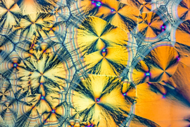 Abstraktes Makrofoto von Vitamin-C-Kristallen unter kreuzpolarisiertem Licht. Die Kristalle erscheinen in leuchtenden Gelb-, Blau- und Rottönen.