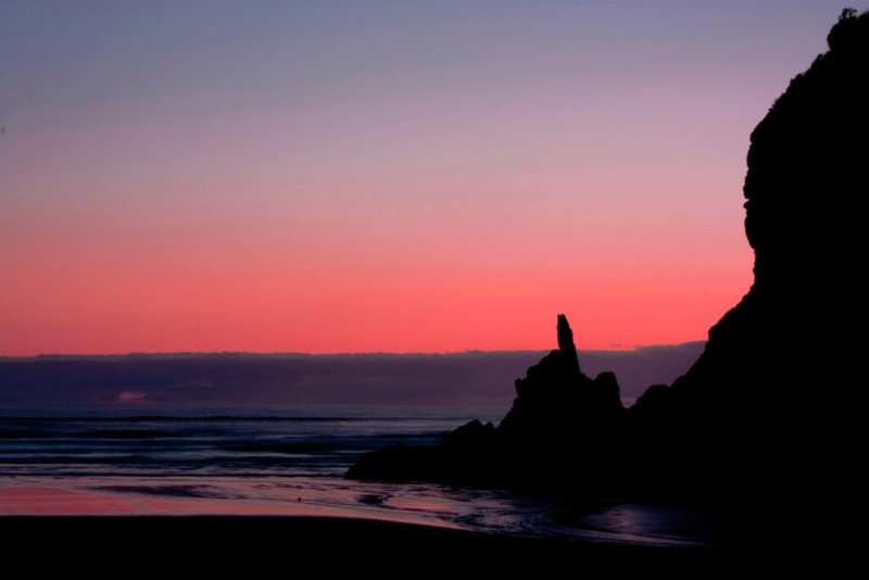 Silhouette eines Felsvorsprungs bei Sonnenuntergang mit einem Himmel in rosa und violetten Tönen und einem ruhigen Meer.